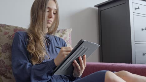 Mujer-Joven-Dibujando-En-Una-Tableta-En-El-Sofá-De-Casa,-Vista-Frontal