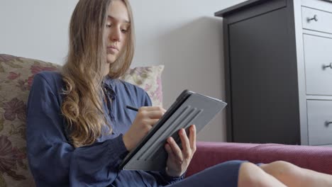 mujer joven dibujando en una tableta en el sofá de la casa, vista frontal
