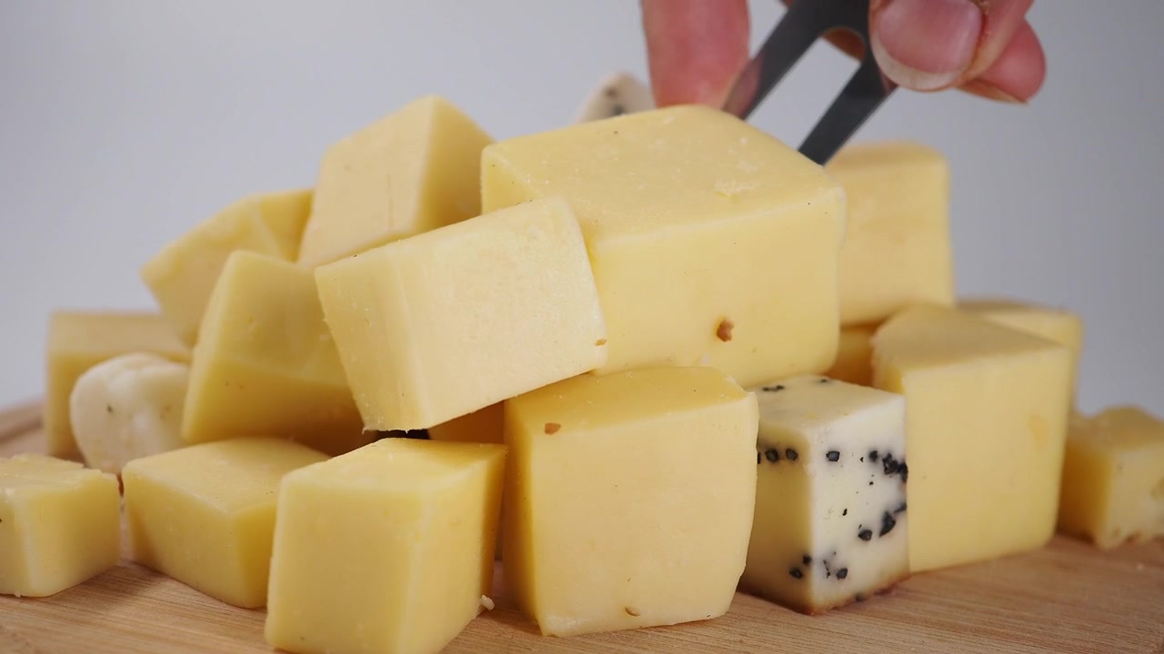 Mano Recogiendo un Cubo de Queso Amarillo de una Pila con Pinzas.