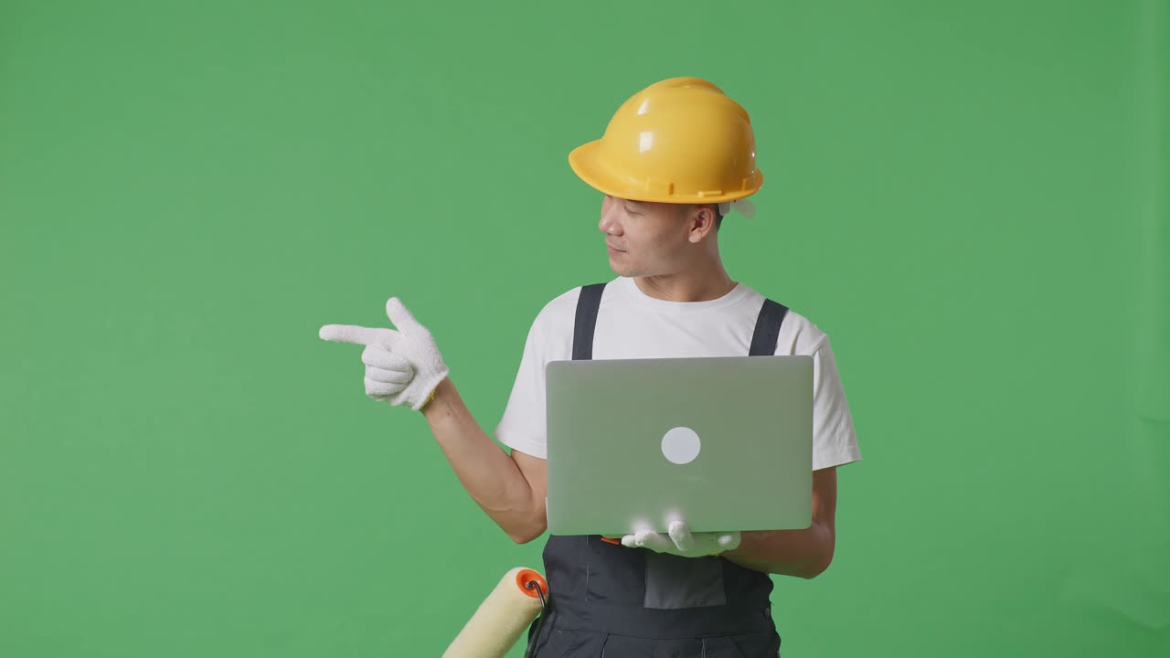Asian Man Painter Wearing Safety Helmet Using A Laptop And Pointing To Side While Standing In The Green Screen Background Studio