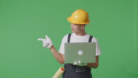 Asian Man Painter Wearing Safety Helmet Using A Laptop And Pointing To Side While Standing In The Green Screen Background Studio
