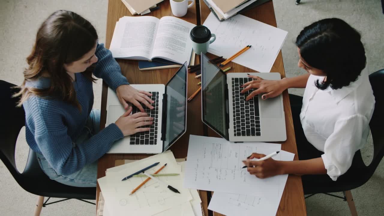 Opening laptops, two women blue-sweater white-top typing and writing at study table, preparing work