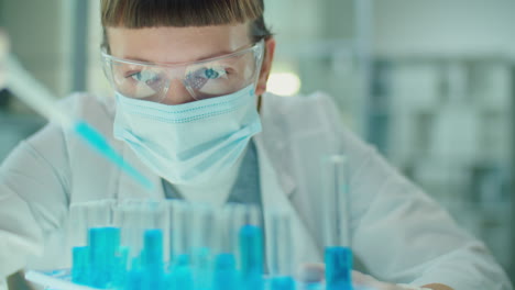 woman in mask and gloves pouring blue chemical in test tubes
