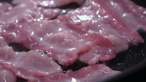 closeup view: pink bacon strips fry, sizzle, bubble in hot frying pan