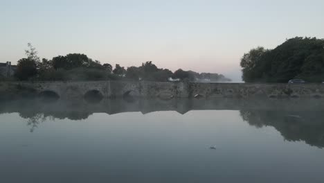 traffic over stone bridge near adare castle during foggy sunrise in county limerick, ireland. aerial dolly shot
