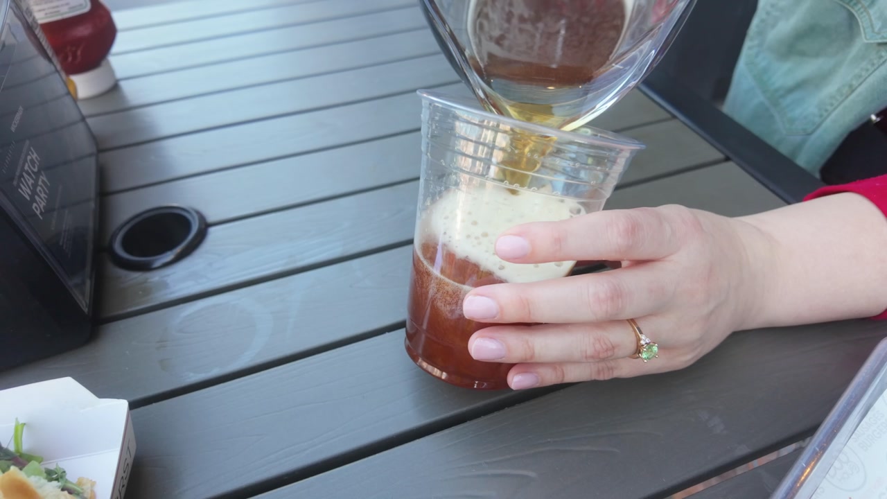 Pouring a beer into a cup from a pitcher at an outdoor table in a slow motion close up.