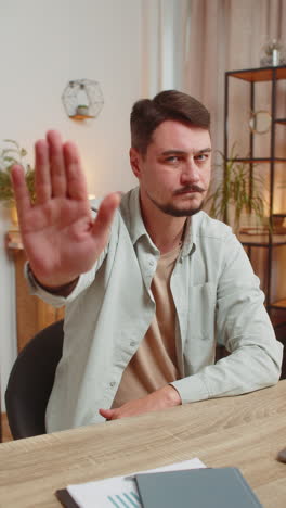 young man freelancer with serious expression doing stop gesture with both hands at home office desk