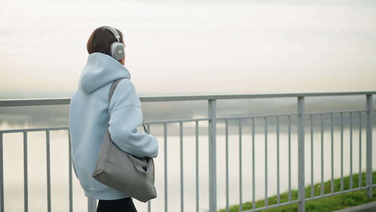 Backside view of female adult walking beside iron rail, holding a bag, with river in the background, the scene evokes tranquility and peacefulness as she strolls along the railing near water