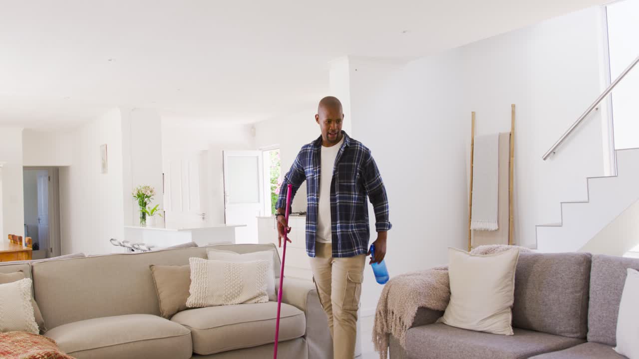 African American man entering living room with pink mop and blue spray bottle, scanning to clean