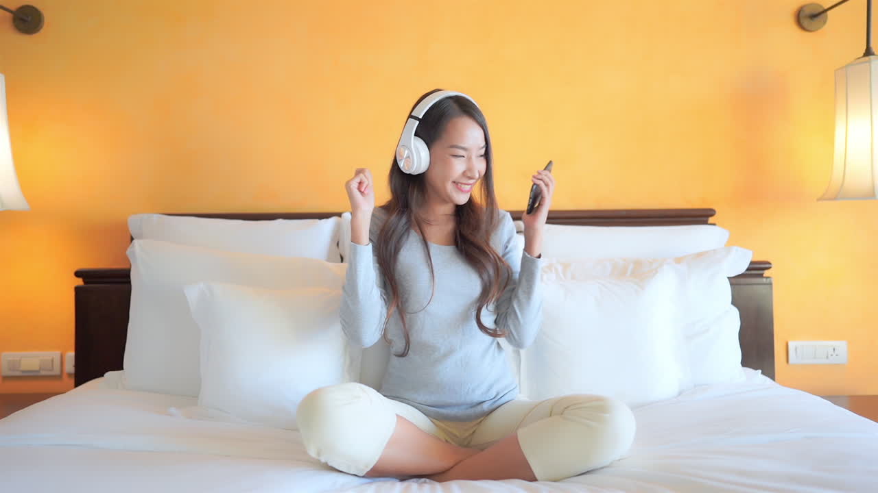 Woman with headphones holding cell phone and dancing to streaming music while sitting on bed. Bright morning sunlight shines into bedroom of woman listening to music seated on bed