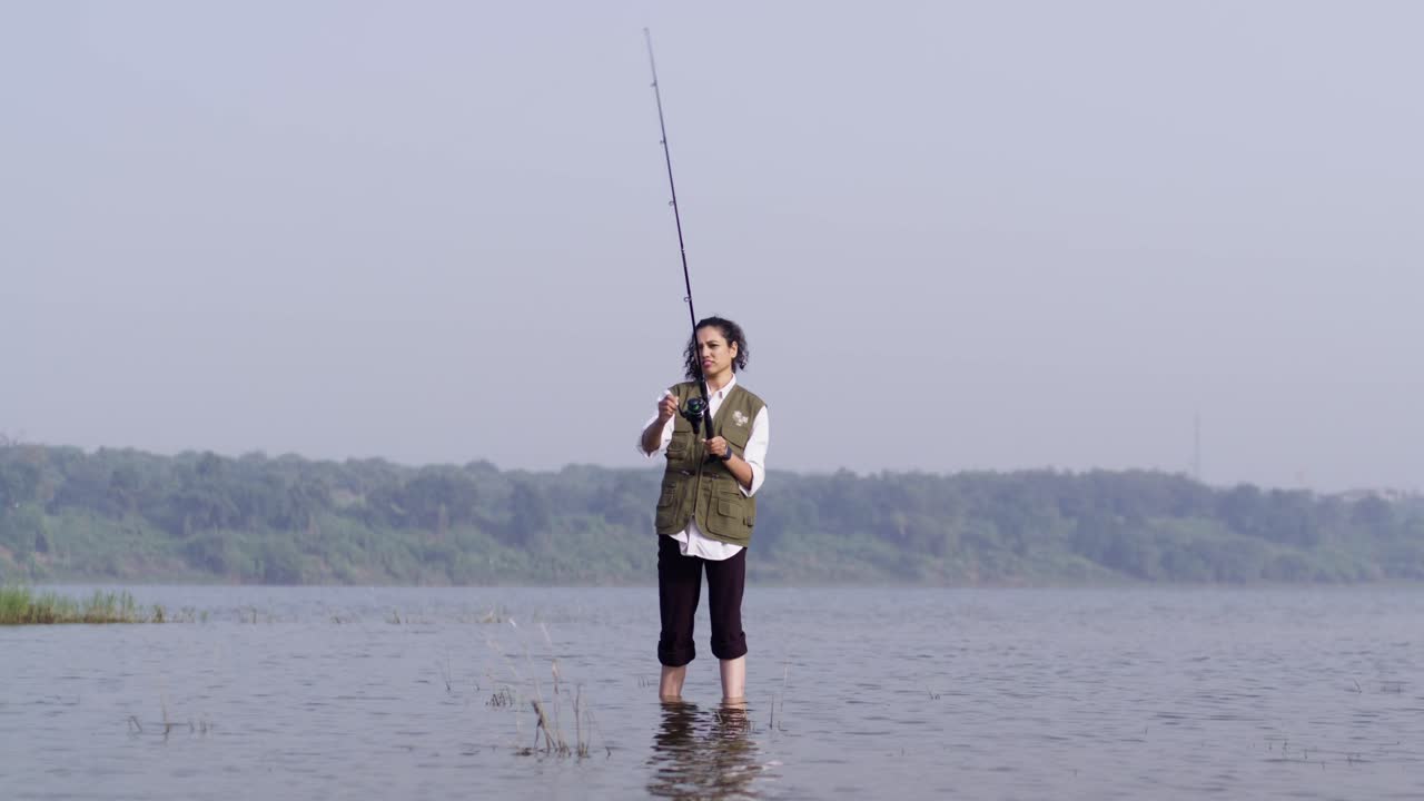 Seorang gadis Asia Selatan mengenakan rompi memancing berdiri di air danau yang dangkal, melemparkan pancingnya ke arah perairan terbuka