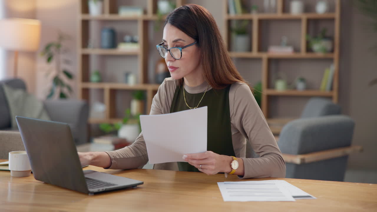 una mujer trabaja en su oficina de casa, escribiendo en una computadora portátil y leyendo un documento.