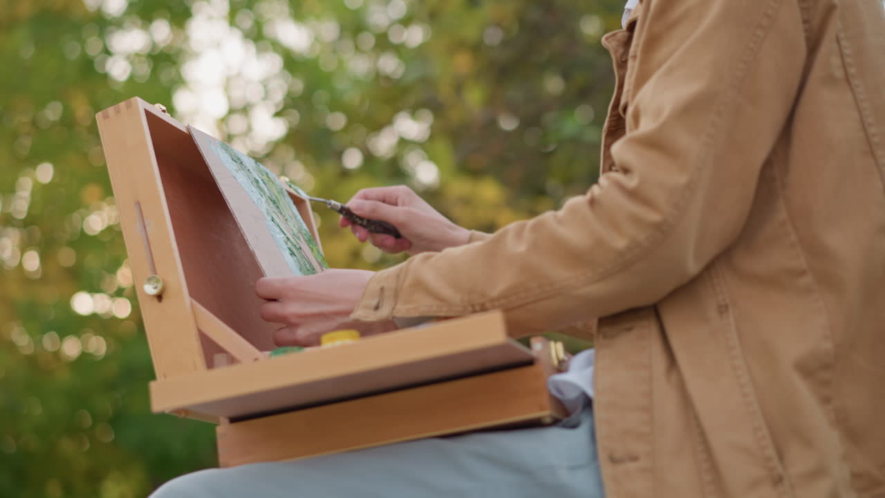 Outdoor painter mixing colors on palette while seated in park closeup of hands applying brush strokes to canvas inside wooden pochade box, soft autumn sunlight, leafy background, relaxed creative