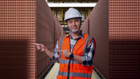 asian male engineer with safety helmet smiling and pointing to side while standing with red brick packed in stacks are stored