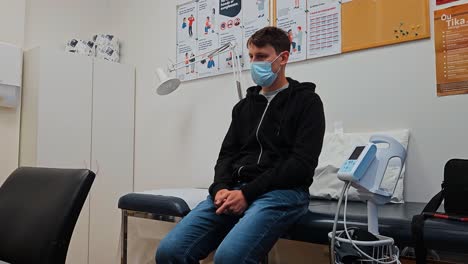 male patient with face mask sitting in hospital exam room and waiting