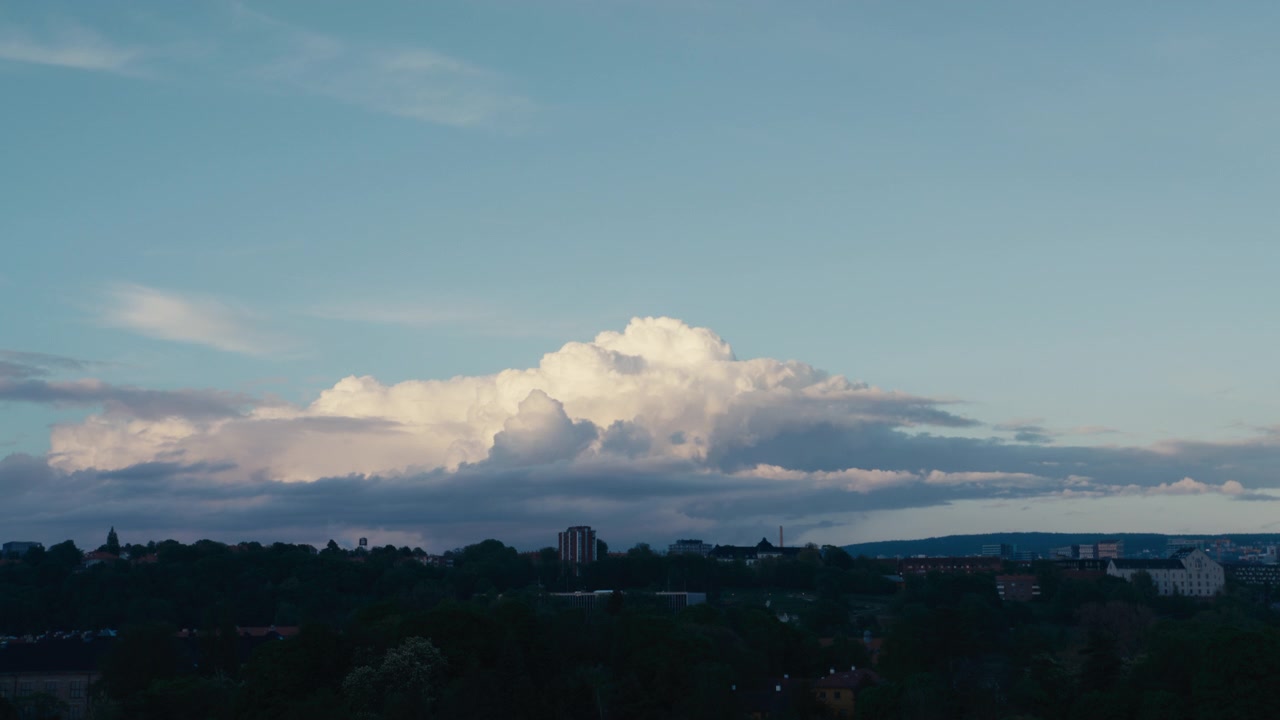 Large cumulus clouds glow in the evening light above a dark Norwegian town skyline with distant hills and soft blue sky.