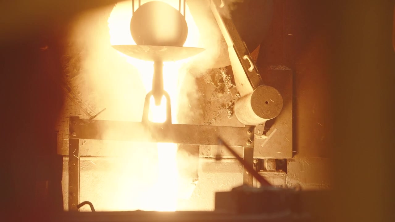 White hot molten metal gushes from a suspended ladle into a glowing chamber, filling the frame with intense light and steam.