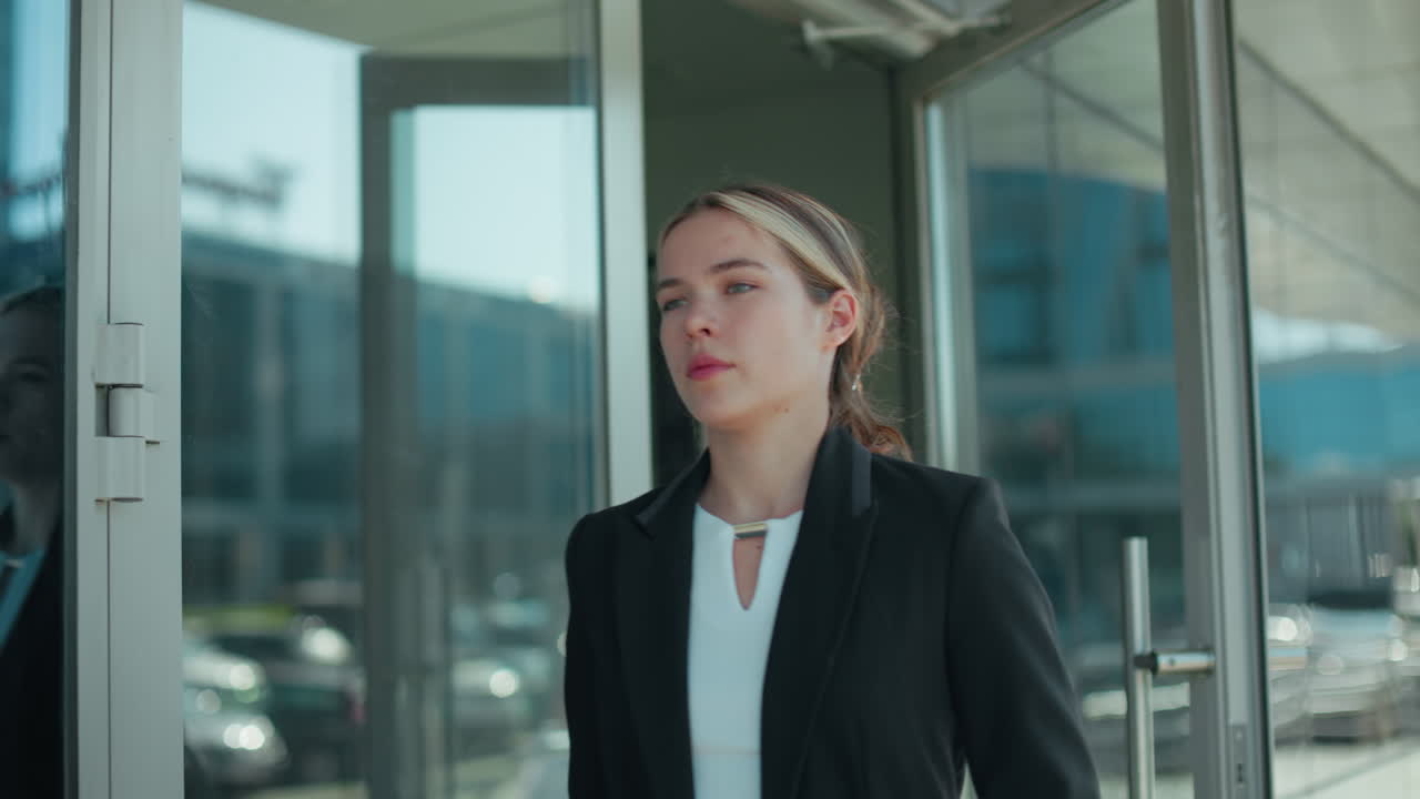 Professional designer opens glass door walking out of building with her reflection visible on door surface, parked cars and trees lining urban street in background