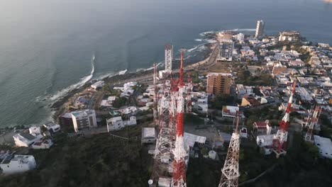 Drohne-Dreht-Sich-Um-Die-Basisstation-Des-Telekommunikationsturms-über-Der-Klippe,-Dem-Meer,-Bei-Sonnenuntergang-Mit-Der-Skyline-Der-Stadt