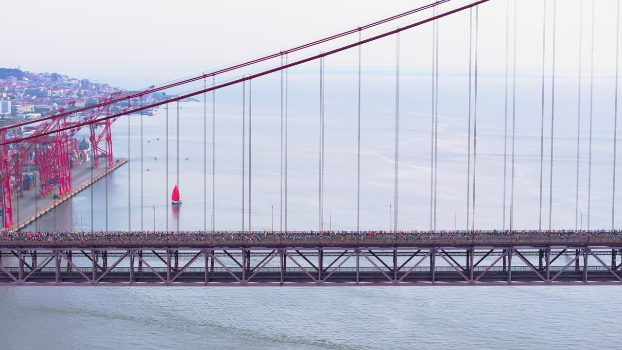 Toma lateral aérea con dron de una media maratón y una carrera de larga distancia de 10 km en Lisboa, Portugal, Europa. Corredores cruzando el puente colgante rojo 25 de Abril. Gradación de color comercial
