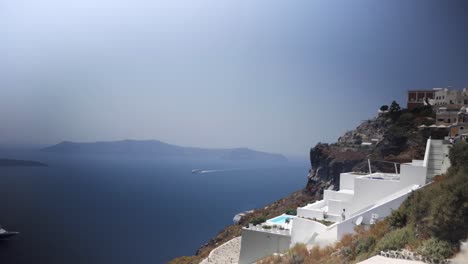 wide shot of a ferry slowly coming into the city of thira in santorini greece, with some islands in the background and a cliff with a white building on it where people are walking