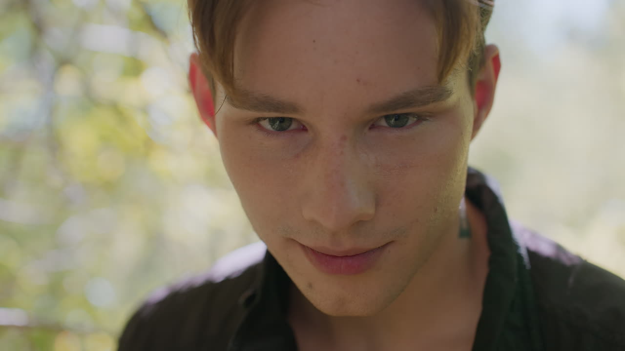Young man lowers head then looks upward with mix of seriousness and slight smile signifying a solution and courage, standing in forest with soft natural lighting highlighting facial features