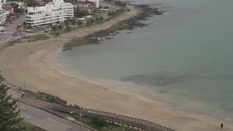 Toma-Aérea-De-Lapso-De-Tiempo-De-Pareja-Y-Perro-Caminando-Por-La-Playa-De-Arena-Durante-El-Día-Nublado-En-Uruguay