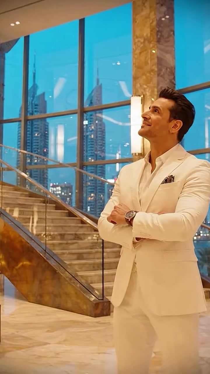Uomo elegante in abito bianco posa in un interno lussuoso con lo skyline della città sullo sfondo al tramonto, mettendo in mostra stile e sicurezza in un contesto di architettura moderna.