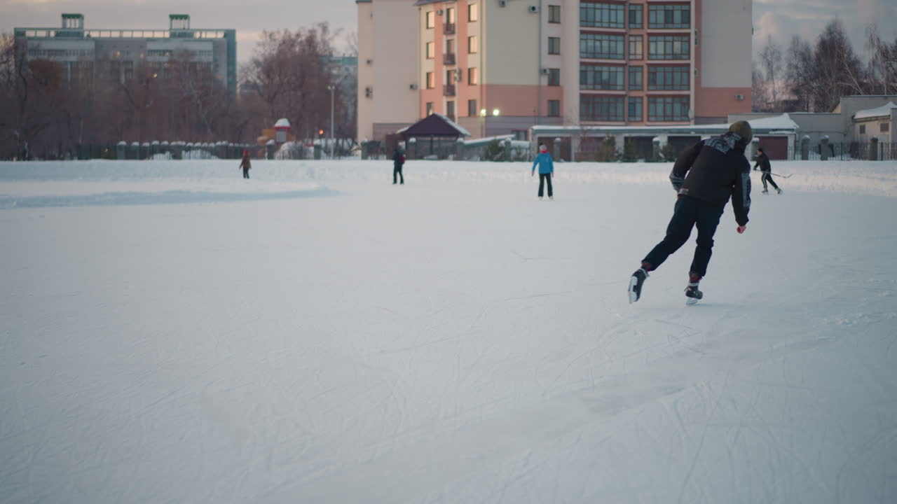 young adult wearing patterned jacket and green hoodie gliding smoothly on frozen lake near urban buildings under pastel sky, people skating in background