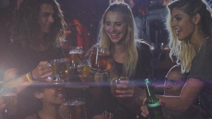 Amici che alzano boccali di birra e inclinano una bottiglia verde per brindare al bar sotto un'illuminazione rosso-blu