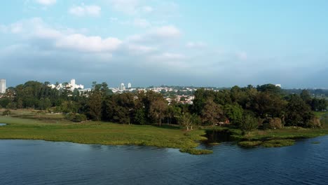 Aerial-drone-dolly-in-rising-shot-of-the-Interlagos-neighborhood-in-the-south-of-São-Paulo,-Brazil-from-the-man-made-Guarapiranga-Reservoir-with-calm-waters,-trees,-and-houses-on-a-fall-evening