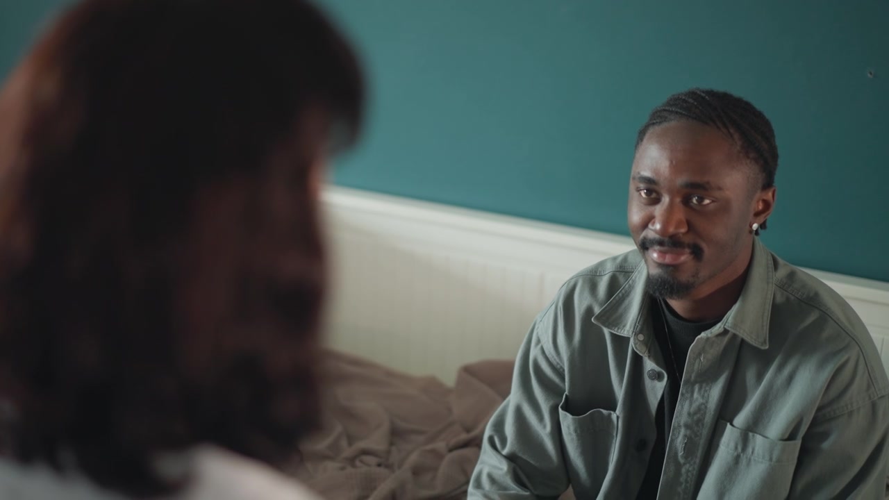 Man smiling during home aftercare visit nurse provides reassurance and gentle bedside check, teal wall bedroom backdrop, warm natural light, relaxed posture, hopeful conversation, recovery progress
