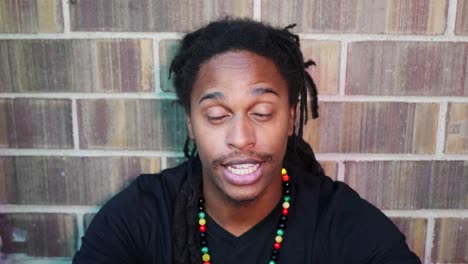 hombre afroamericano con rastas sentado en el suelo mirando a la cámara diciendo &quot;no&quot;, &quot;sí&quot; y &quot;tal vez&quot;, sacudiendo la cabeza y sonriendo