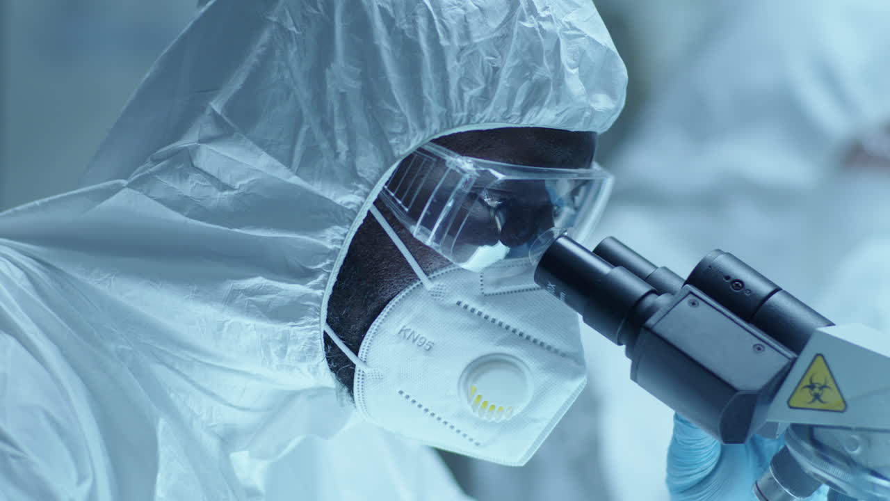Chemist in Protective Gear Using Microscope during Medical Test in Laboratory