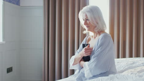 Bereaved-Senior-Woman-Sitting-On-Edge-Of-Bed-Looking-At-Photo-In-Frame