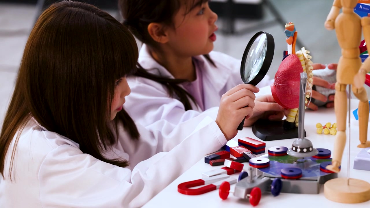 Two young students in white coats use a magnifying glass to study a lung model.
