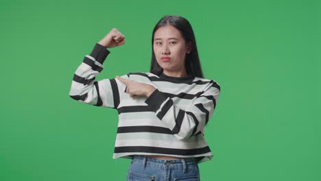 a smiling asian woman pointing bicep while standing in front of green screen background