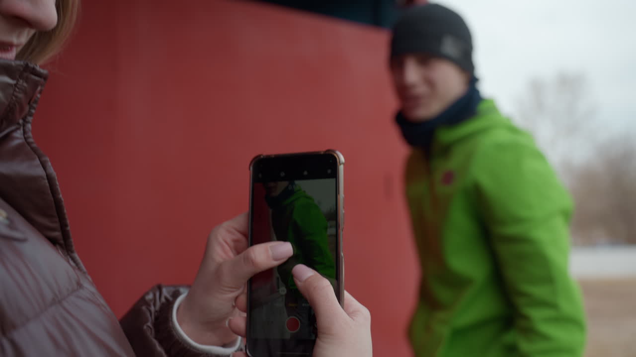 Una mujer hace una foto en el exterior, un fotógrafo con un teléfono saca una foto en un parque durante el invierno, una mujer con un abrigo de invierno hace un retrato espontáneo en el exterior con su dispositivo móvil para compartirlo en redes sociales.