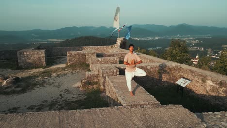 orbita cámara lenta de hombre indio haciendo hatha yoga de pie en una pierna en la pared del castillo de piedra en la cima de la colina rodeado de colinas campos y bosques por la mañana al amanecer
