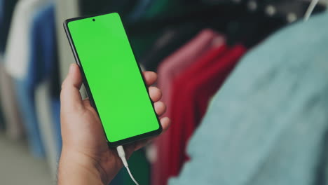 un hombre con camisa verde azulado sostiene firmemente su teléfono móvil mientras mira y escucha a través de los auriculares en una tienda de ropa.