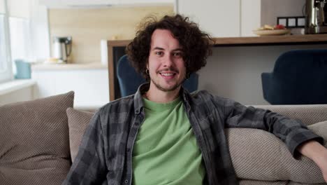 Portrait-of-a-happy-brunette-guy-with-curly-hair-a-mustache-and-a-plaid-shirt-who-sits-on-the-sofa-in-a-modern-studio-apartment