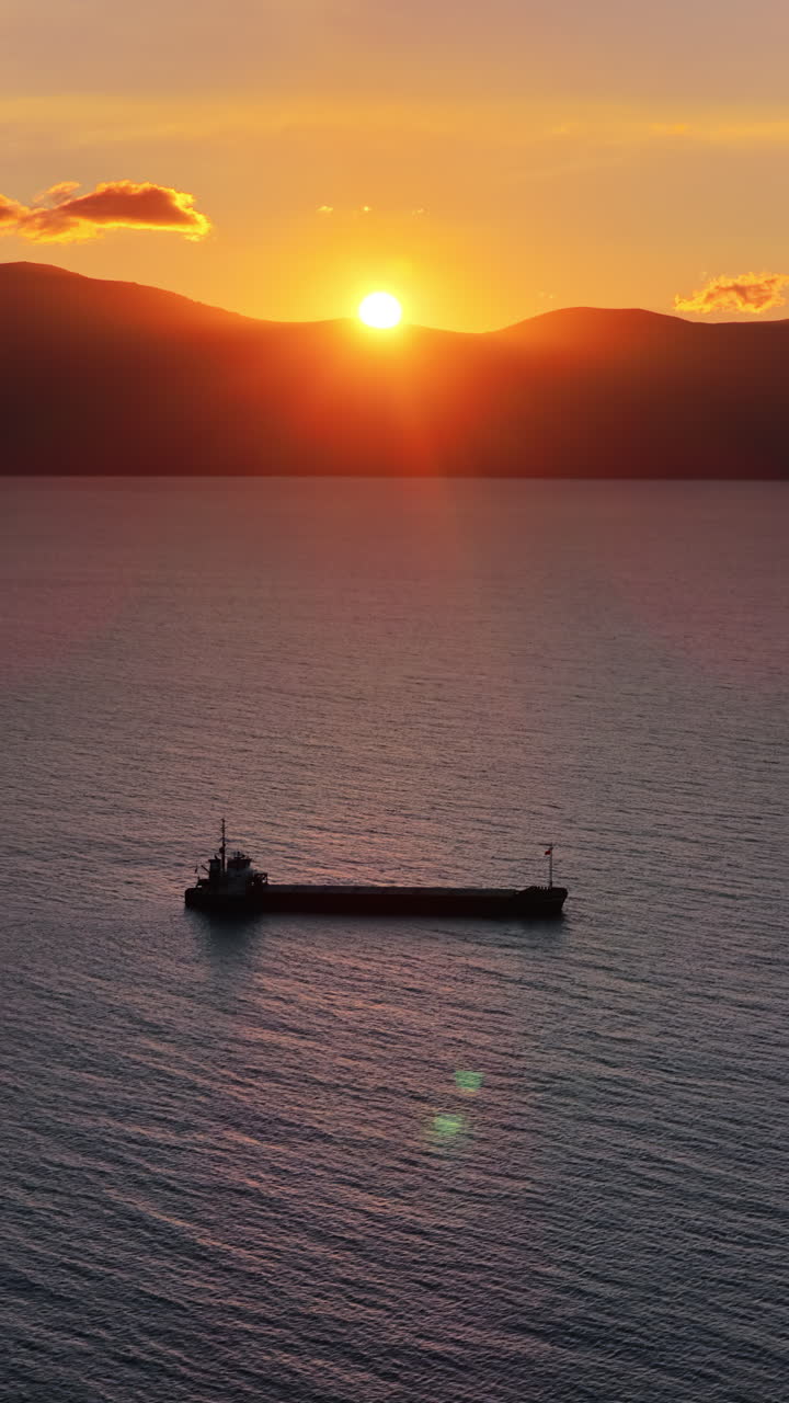 Aerial drone view of a fisherman rowing a weathered wooden boat across the serene waters of Vlora Bay at dusk, creating gentle ripples that reflect the pastel evening light. Vertical