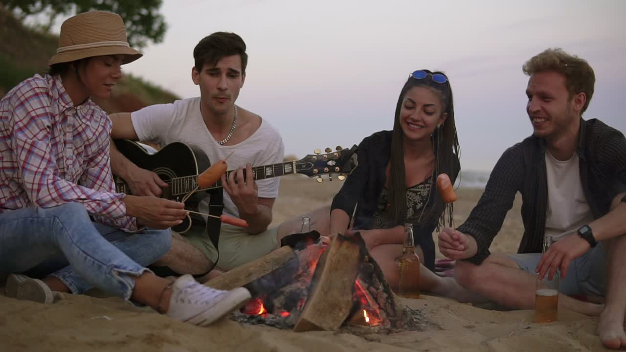 grupo de jóvenes y alegres sentados junto al fuego en la playa por la noche, asando salchichas y tocando la guitarra