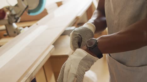 Mid-section-of-african-american-male-carpenter-wearing-safety-gloves-in-a-carpentry-shop