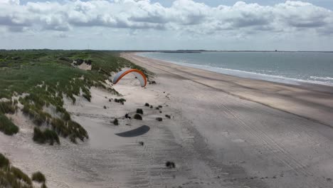 Paraglider-landing-on-empty-beach-by-lush-green-waterfront-sand-dunes