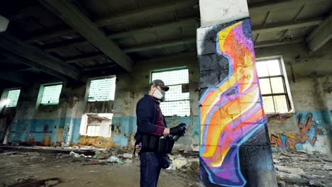 low angle shot of young man is face mask painting graffiti on column inside empty industrial building using spray paint. dirty damaged walls, windows and ceiling are visible.