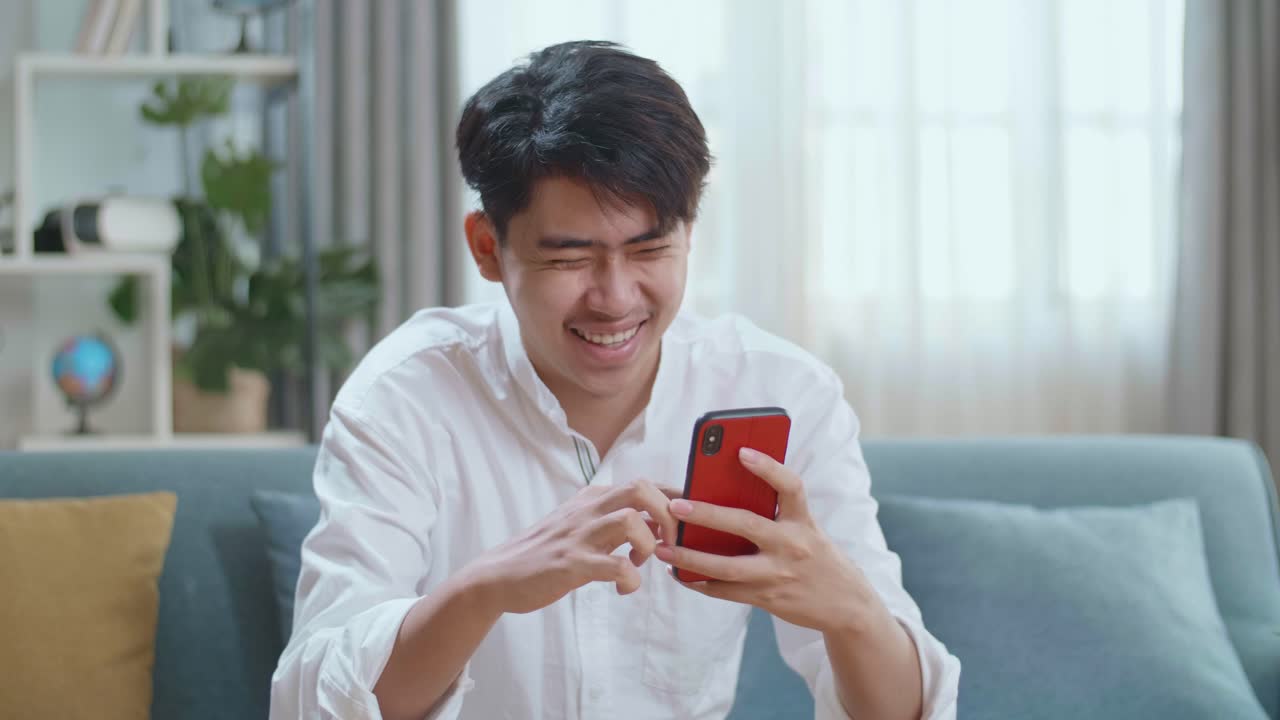Asian Man Touching Smartphone And Laughing While Sitting On Sofa In The Living Room