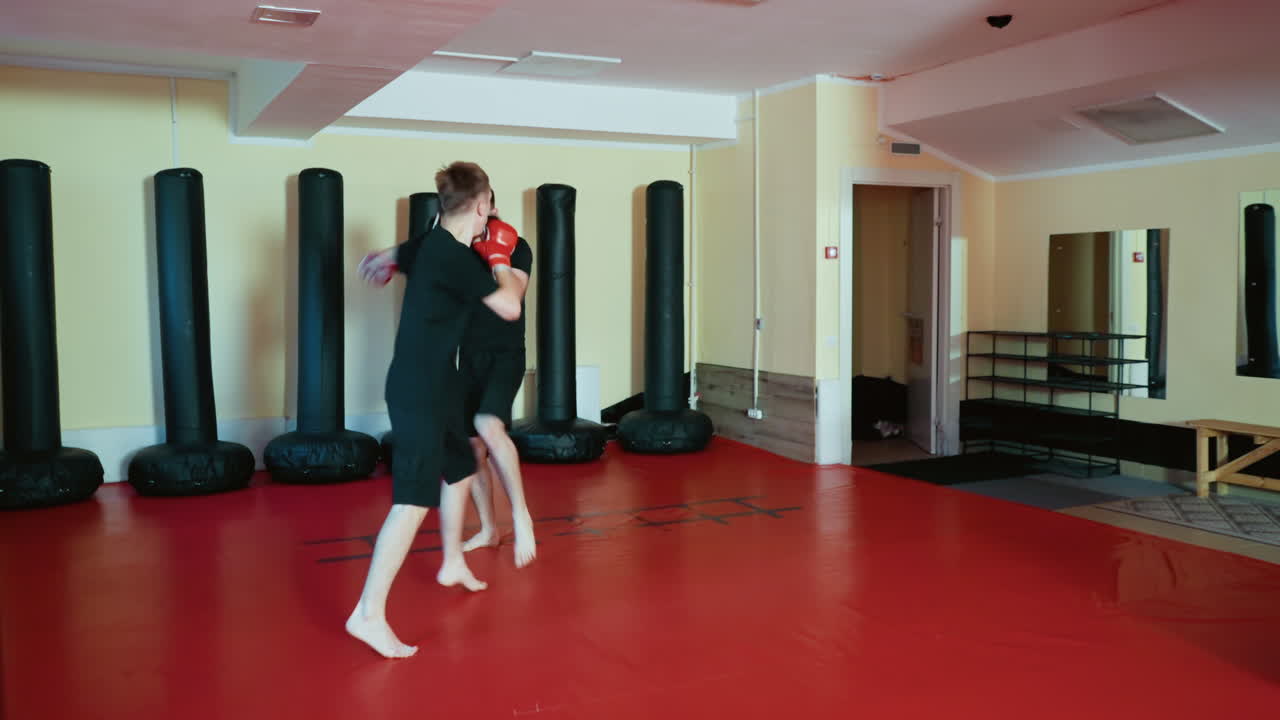 Wrestlers sparring inside gym, wearing gloves and black outfits, practicing combat techniques with striking moves and defensive stance on red mat floor near punching bags during intense martial training