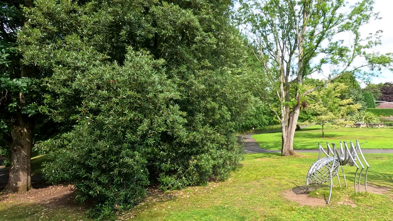 Camera pans right across a sunlit botanical garden, revealing large metallic insect sculptures among lush green trees and manicured lawns under natural daylight
