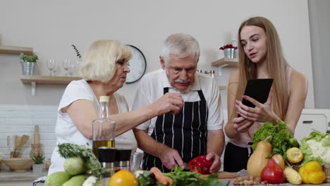 Pareja-De-Abuelos-Maduros-Cortando-Verduras-Para-Ensalada,-Escuchando-La-Receta-De-Una-Chica-Con-Tableta